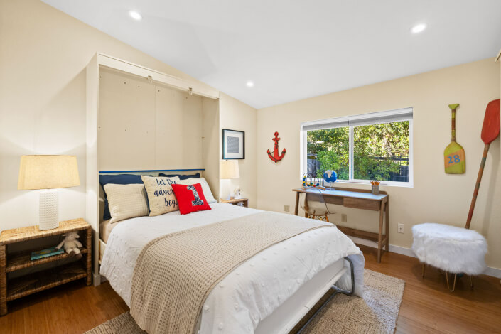 A cozy bedroom with a bed, nautical-themed pillows, a nightstand with a lamp and dollhouse, a desk with a globe and toys, a clear chair, framed art, and a red anchor wall decoration.