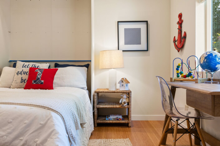 A cozy bedroom with a bed, nautical-themed pillows, a nightstand with a lamp and dollhouse, a desk with a globe and toys, a clear chair, framed art, and a red anchor wall decoration.