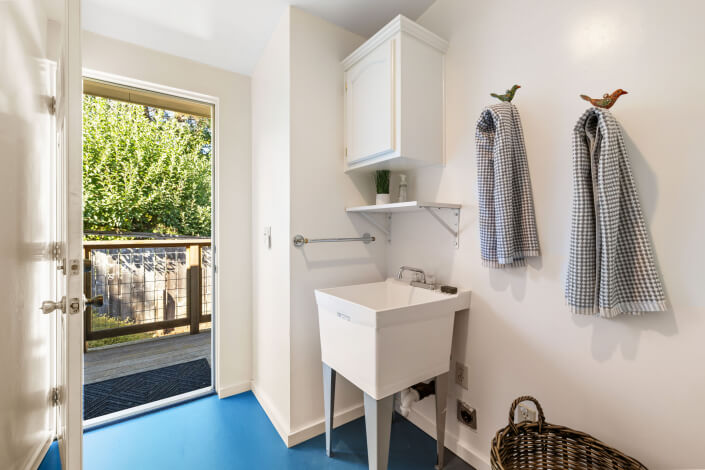 A bright laundry room with blue flooring, a white utility sink, checked hand towels on hooks, wicker laundry basket, cabinet, and an open door leading to a sunny deck and green trees outside.