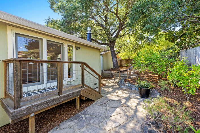 Small patio with stone walkway, wooden railing, and steps leading to a house with large glass doors. Surrounded by mature trees, greenery, and a wooden fence in a shaded backyard.