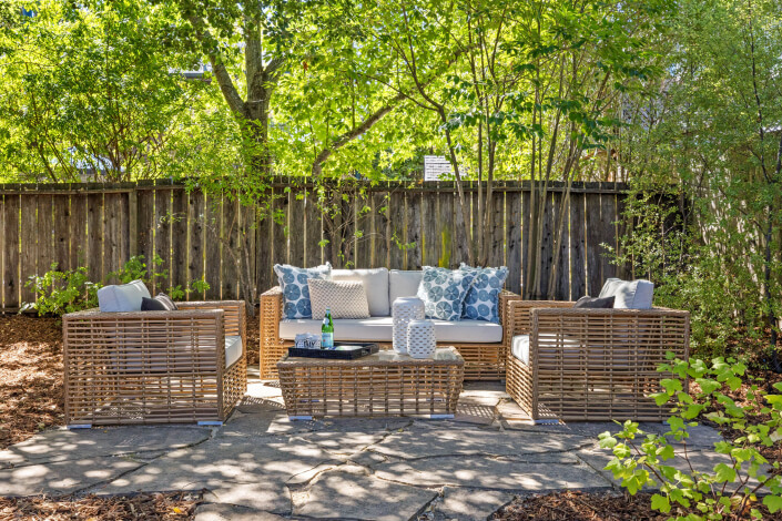 An inviting outdoor patio area with wicker furniture, including a sofa, two chairs, and a coffee table, decorated with blue and white pillows, set on stone pavers and surrounded by lush greenery and trees.