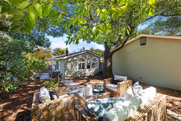 Outdoor seating area with wicker furniture and cushions under large leafy trees, set on a stone patio beside a light green house with arched windows on a sunny day.
