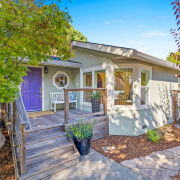 A charming house at 102 Clorinda Avenue in Gerstle Park, San Rafael, features a light gray exterior, a purple front door, white porch bench, wooden steps, potted plants, and trees under a clear blue sky.