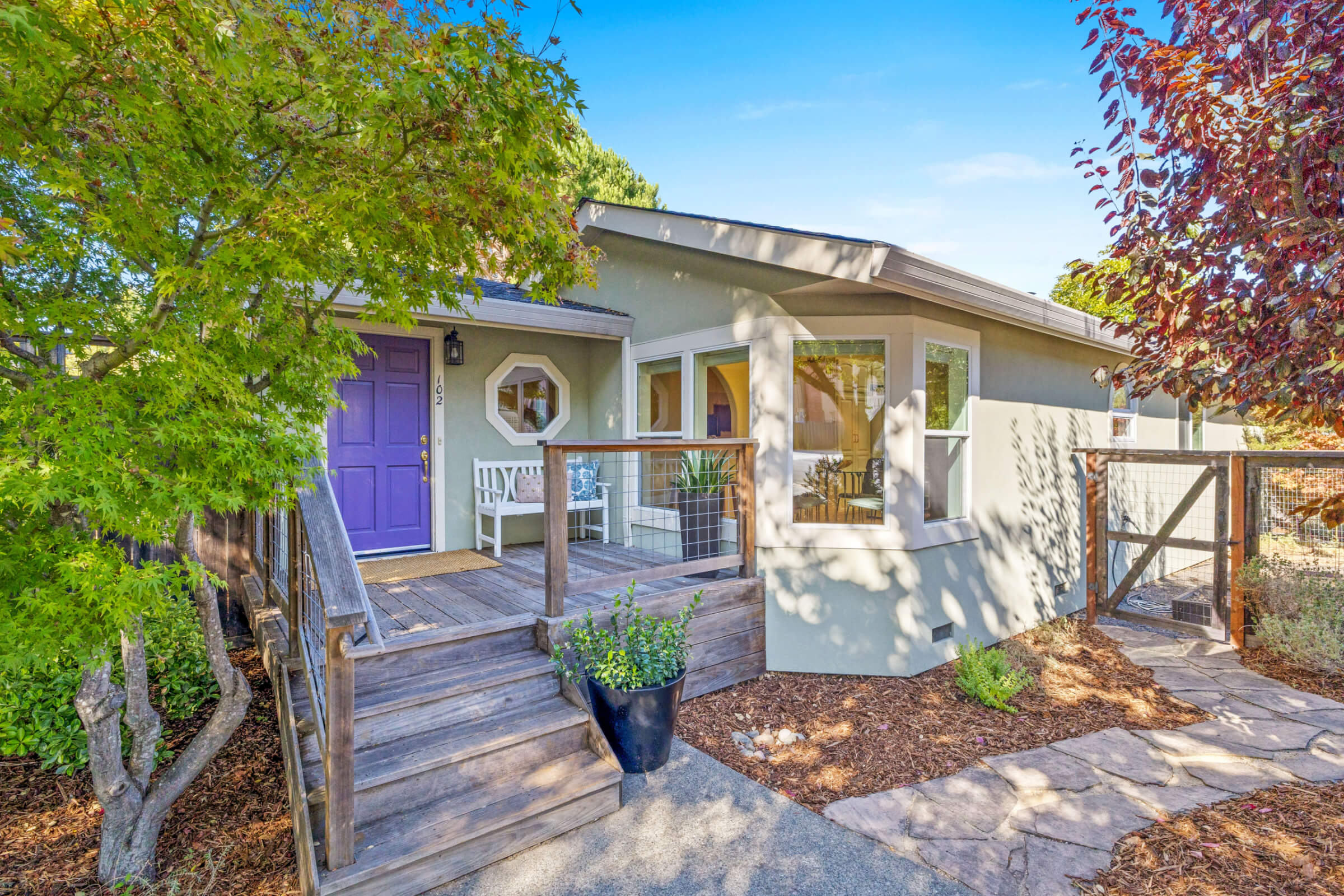 A charming house at 102 Clorinda Avenue in Gerstle Park, San Rafael, features a light gray exterior, a purple front door, white porch bench, wooden steps, potted plants, and trees under a clear blue sky.