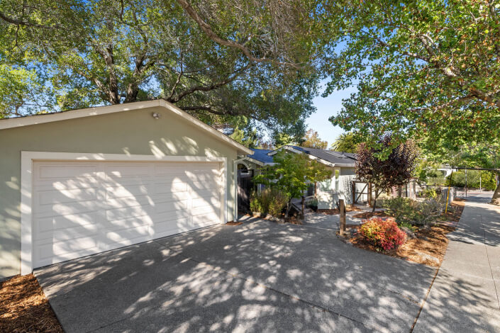 Single-story house with a white garage door, light exterior, and shaded driveway surrounded by trees and landscaped plants on a sunny day.
