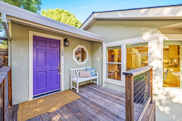A charming house at 102 Clorinda Avenue in Gerstle Park, San Rafael, features a light gray exterior, a purple front door, white porch bench, wooden steps, potted plants, and trees under a clear blue sky.