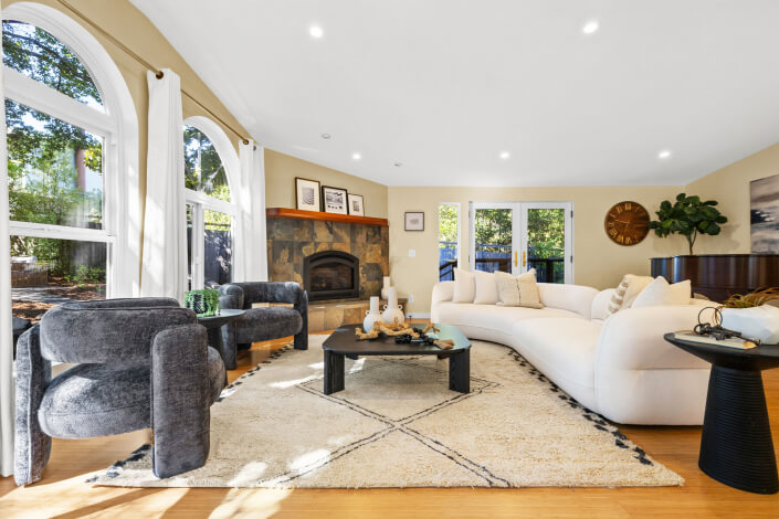 A bright living room at 102 Clorinda Avenue Gerstle Park San Rafael features large arched windows, a stone fireplace, a white sectional, gray chairs, a light rug, black coffee table with decor, and wood floors. Trees are visible outside.