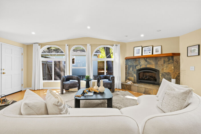 A bright living room with large arched windows, two dark armchairs, a curved white sofa, a black coffee table, and a stone fireplace with artwork on the mantel. Sunlight streams in, creating a cozy atmosphere.