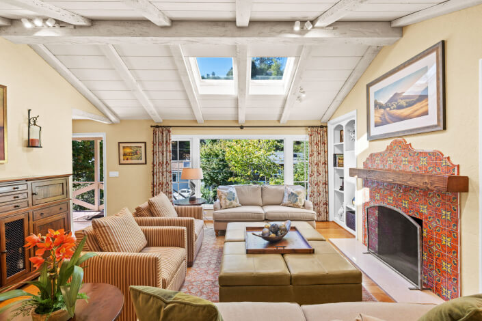 A bright living room with a beamed ceiling, skylights, beige sofas, patterned armchairs, a tiled fireplace, and large windows overlooking green trees. A bowl decorates a central ottoman, and artwork hangs above the fireplace.