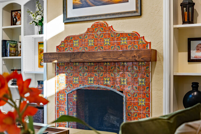 A fireplace with a decorative wooden mantel and vibrant orange patterned tiles, surrounded by cream walls and built-in shelves with books, photos, and decor items. A framed painting hangs above the fireplace.