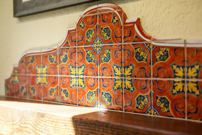 Colorful, ornate ceramic tiles with intricate patterns in orange, blue, and yellow form a decorative backsplash above a wooden surface against a textured beige wall. Sunlight casts shadows on the tiles and wood.