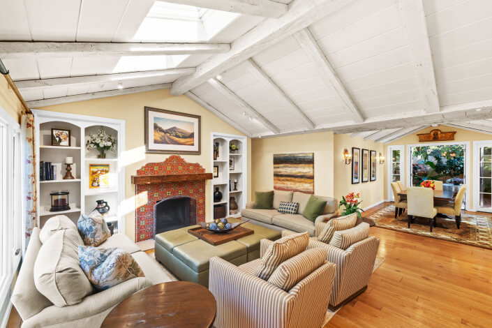 Bright living room at 260 Clorinda Avenue San Rafael with a white vaulted ceiling, exposed beams, colorful tiled fireplace, beige sofas, and striped armchairs. Large windows and glass doors fill the space with natural light; dining area in view.