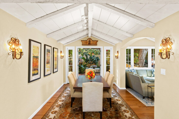 A bright dining room with a vaulted white ceiling, beige walls, framed art, and two ornate wall sconces. A wooden table with six upholstered chairs sits on a patterned rug; large windows overlook a garden. A living room is visible to the right.