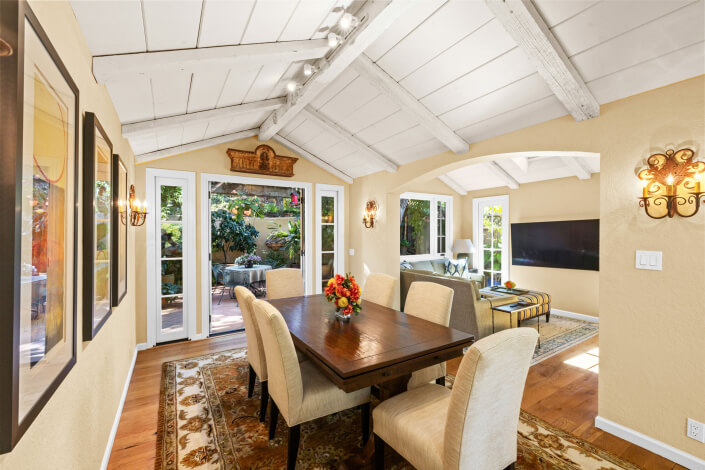 A bright dining room at 260 Clorinda Avenue San Rafael features a wooden table, six upholstered chairs, a floral centerpiece, large windows and glass doors to the garden, decorative wall sconces, and a living area with a TV.