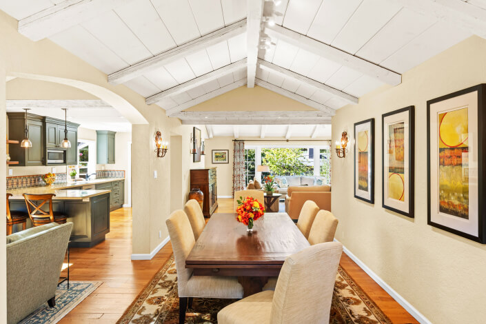 Bright dining room at 260 Clorinda Avenue San Rafael with vaulted white ceiling, wooden table and six beige chairs, floral centerpiece, three colorful framed artworks, and open view to kitchen and living room with large windows.