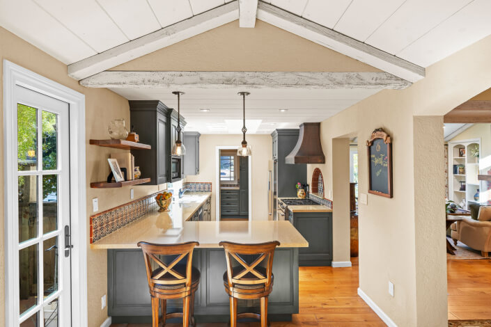 Bright kitchen with exposed beams, pendant lights, a central island with two wooden chairs, dark cabinets, granite countertops, and wood floors. French doors and a living room are visible in the background.