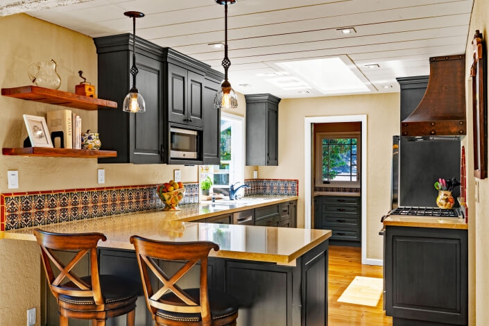 A stylish kitchen with dark cabinets, tan countertops, patterned tile backsplash, wooden shelves with decor, two wooden barstools, pendant lights, and a window letting in natural light.