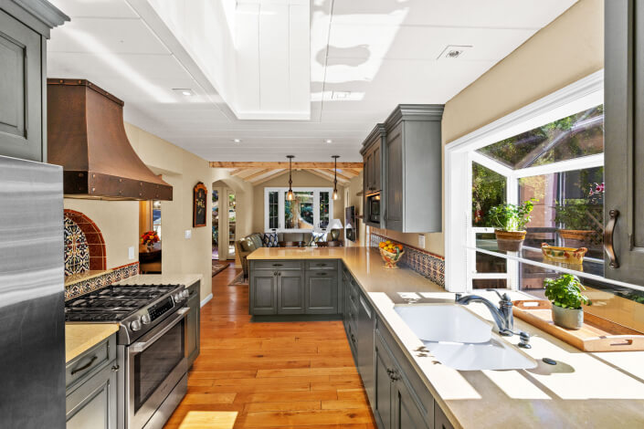 Bright kitchen with stainless steel appliances, dark cabinets, a farmhouse sink, wood floors, and large windows; an open layout leads to a cozy living area with natural light and garden views.
