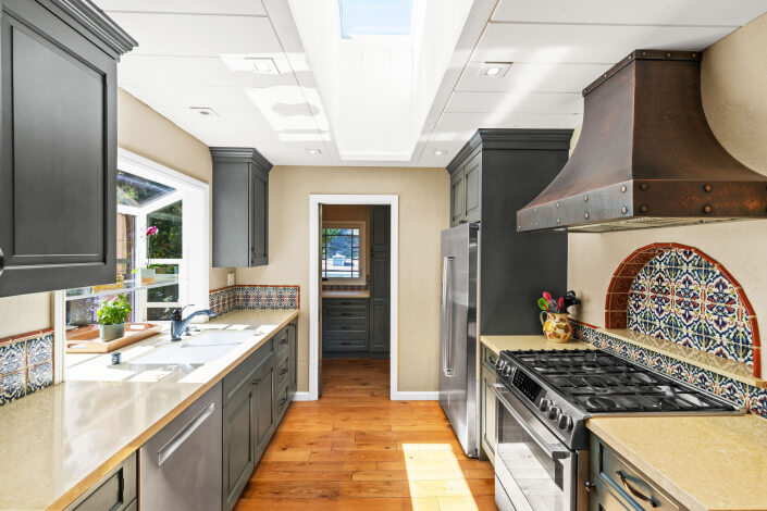 A bright, modern kitchen with dark cabinets, light countertops, a large stainless steel stove and fridge, a decorative tile backsplash, a bronze range hood, wood floors, and sunlight streaming through a skylight.