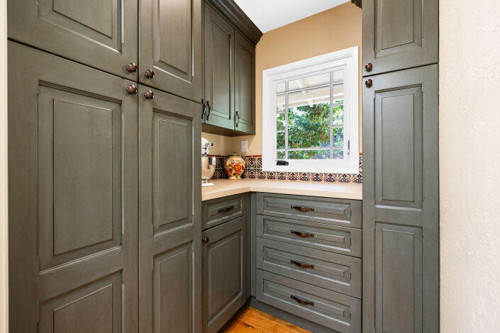 A kitchen with olive green cabinets, brown knobs, beige countertops, a window with white trim, decorative tile backsplash, and wooden floor. A coffee maker and decorative items sit on the counter.