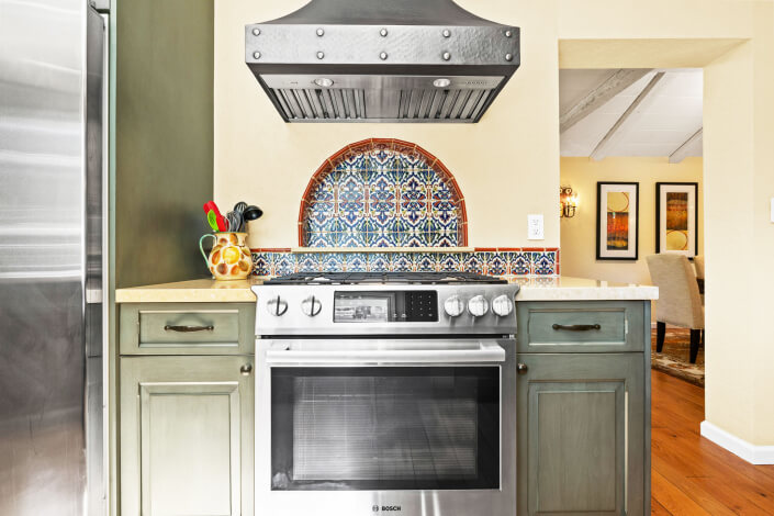 A modern kitchen features a stainless steel Bosch oven, green cabinets, a patterned tile backsplash, a metal range hood, and a vase with utensils on a wooden countertop. Dining area is visible in the background.