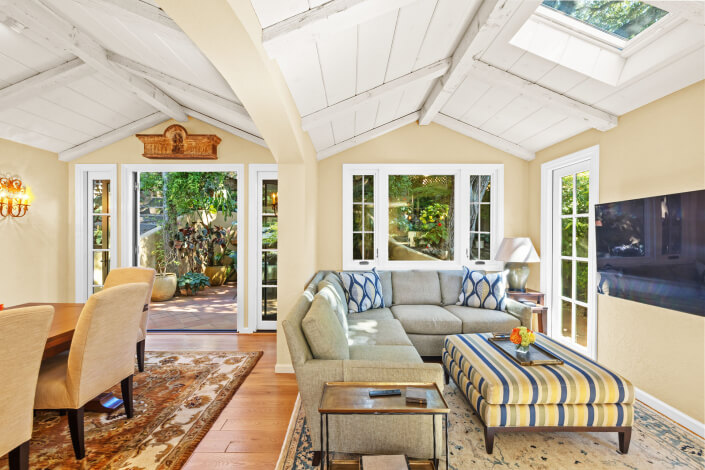Bright living and dining room at 260 Clorinda Avenue San Rafael with vaulted white ceiling, beige walls, a gray sectional sofa, striped ottoman, patterned rug, artwork, and large windows letting in natural light.