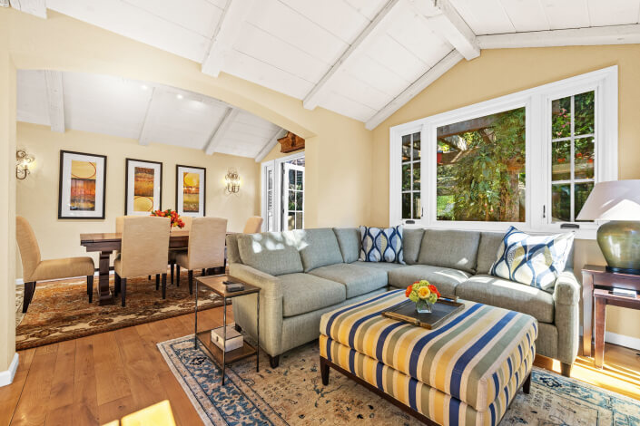 Bright living and dining room at 260 Clorinda Avenue San Rafael with vaulted white ceiling, beige walls, a gray sectional sofa, striped ottoman, patterned rug, artwork, and large windows letting in natural light.