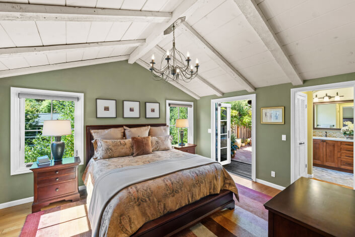 A primary bedroom with green walls, slanted white wooden ceiling, chandelier, wall-mounted TV, wooden dresser, and bed with patterned bedding. French doors open to a sunny patio with outdoor seating.