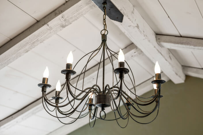 A black metal chandelier with eight lit candle-shaped bulbs hangs from a white wooden ceiling with exposed beams.