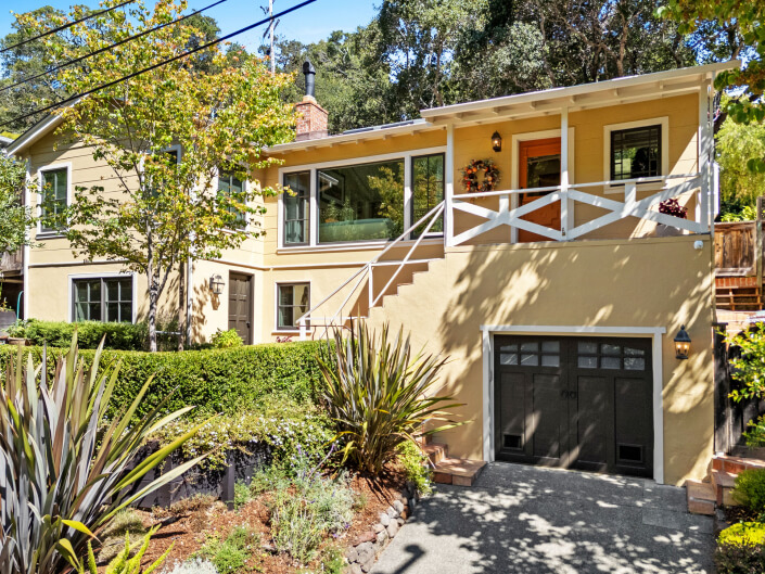 A two-story beige house at 260 Clorinda Avenue San Rafael features large windows, a front porch with white railings, a single-car garage, and a landscaped garden, all surrounded by trees and greenery.