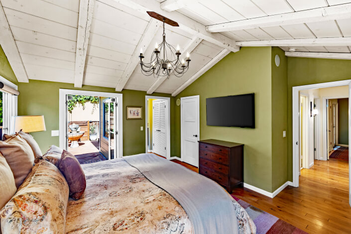 A primary bedroom with green walls, slanted white wooden ceiling, chandelier, wall-mounted TV, wooden dresser, and bed with patterned bedding. French doors open to a sunny patio with outdoor seating.