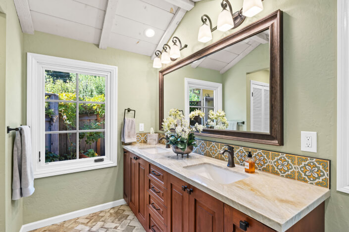 A bright bathroom with a double sink vanity, wooden cabinets, a large framed mirror, decorative tile backsplash, flowers, and a window with a garden view. Towel, soap, and lights are also visible.