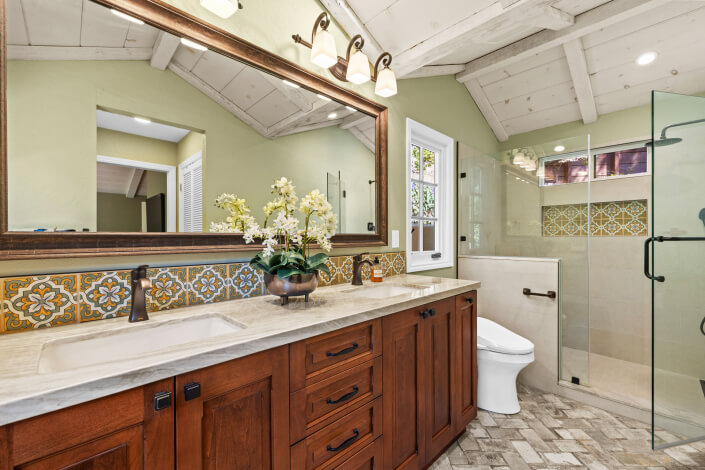 A bathroom with wood cabinets, a marble countertop, wall-mounted lights, a large mirror, patterned tile backsplash, potted flowers, and a glass shower next to a window. The ceiling has exposed white beams.