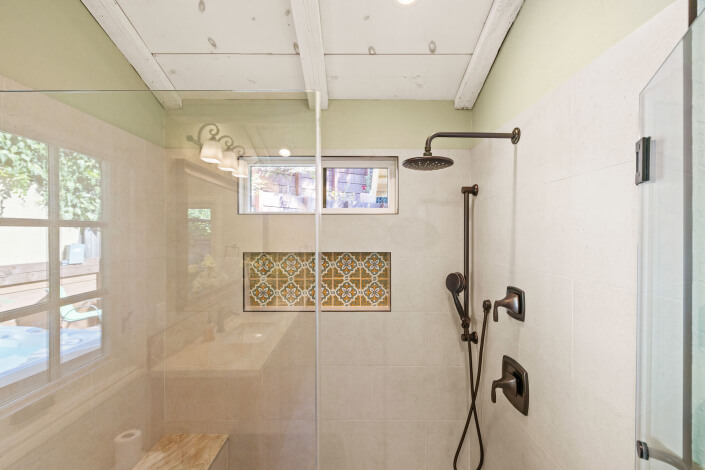 A modern shower with beige tiles, a glass door, a rainfall showerhead, a handheld shower, and a built-in niche with decorative patterned tiles; a small window and wood bench are also visible.