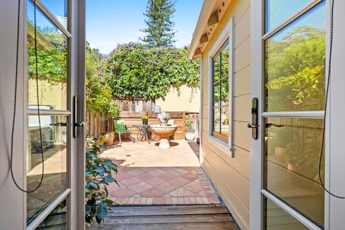 View from inside through open double doors to a sunny patio with terracotta tiles, outdoor seating, a round table, potted plants, and lush greenery against a wooden fence and clear sky.