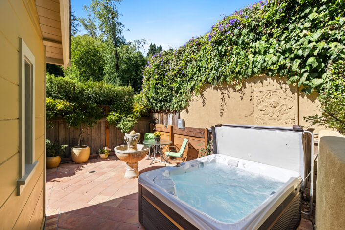 A cozy outdoor patio at 260 Clorinda Avenue San Rafael features a bubbling hot tub, small round table with two chairs, potted plants, and a decorative fountain. Lush greenery and flowering vines cover the fence and walls.
