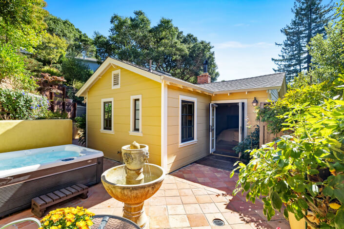 A yellow house with white trim at 260 Clorinda Avenue San Rafael features an open door leading into a bedroom. A jacuzzi, stone fountain, and plants sit on a sunlit tiled patio surrounded by greenery and tall trees.
