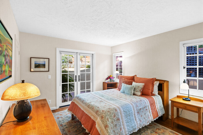 A bedroom with a double bed in peach and blue bedding, two wooden nightstands, a lamp, wall art, and large French doors with a view of a garden. Light streams in through the windows, brightening the room.