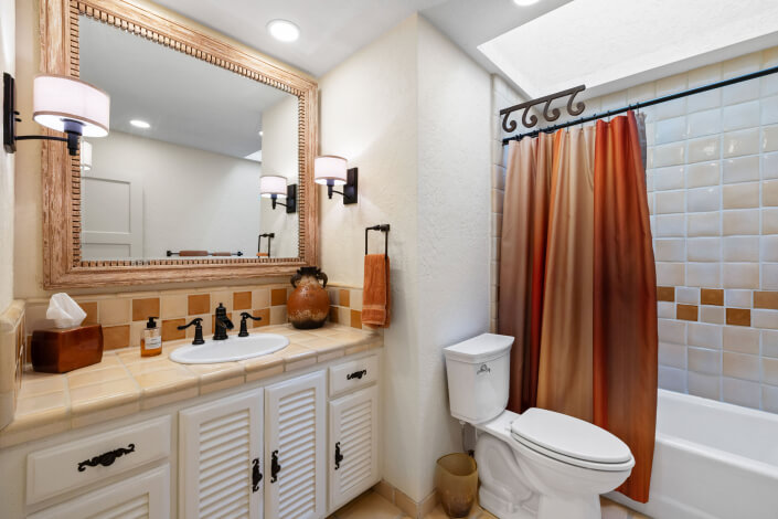 A bathroom with a white sink and cabinets, a large framed mirror, wall sconces, a toilet, and a bathtub with a brown and orange shower curtain. The walls are cream-colored with tile accents.