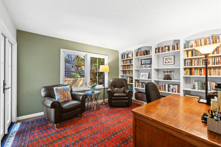 A home office with two leather armchairs, a wooden desk, built-in bookshelves filled with books, a colorful rug, and large windows letting in natural light.