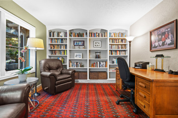 home office with two brown leather chairs, a large wooden desk, bookshelves filled with books, a red patterned rug, and two lamps. A large window lets in natural light, and a family photo hangs on the wall.