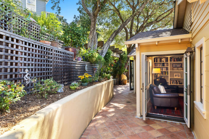 A outdoor patio with terracotta tiles, a garden bed lined with plants and lattice fencing on the left, and an open doorway leading to a book-filled living room on the right. Tall trees provide shade overhead.