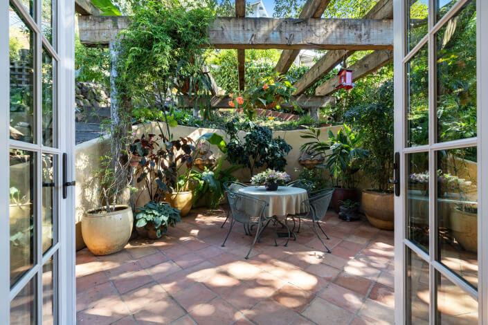 A cozy patio with terra cotta tiles, potted plants, a round table with two chairs, and wooden beams overhead, viewed through open double doors. Sunlight filters through the greenery, creating a tranquil outdoor space.