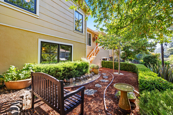 A backyard garden at 260 Clorinda Avenue San Rafael features a wooden bench, birdbath, lush plants, and stepping stones beside a yellow house with large windows. Sunlight filters through the trees onto the inviting space.