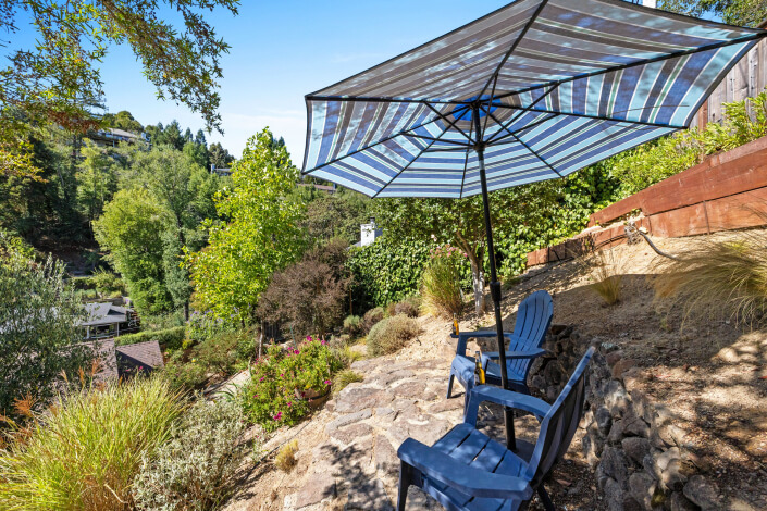 Two blue Adirondack chairs sit on a stone patio under a large striped umbrella, overlooking a lush green hillside with trees and nearby houses on a bright sunny day.