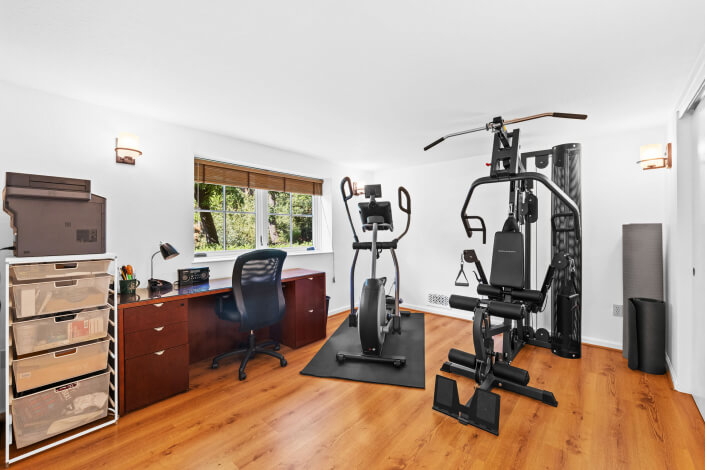 Bright home office and gym with a desk, office chair, printer, and drawers on one side, and exercise equipment including a stationary bike and weight machine on the other, with wood flooring and a window letting in natural light.