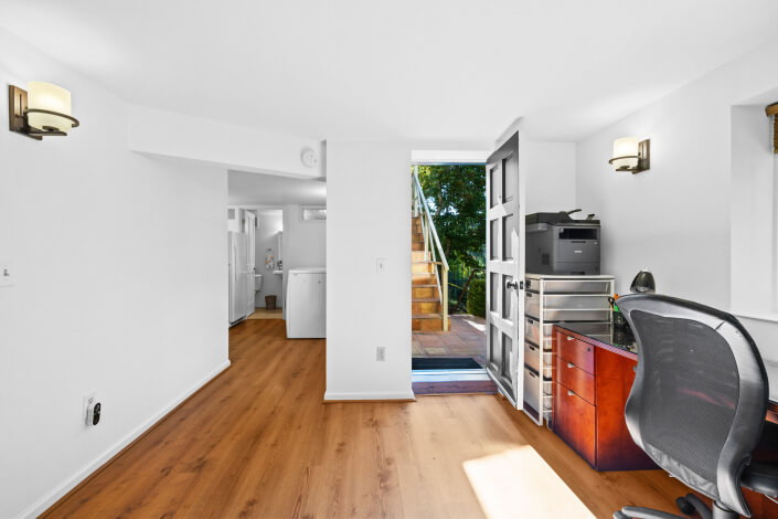 A bright home office with a wooden desk, office chair, and printer sits near an open door leading outside to sunlit stairs, with a laundry area visible in the adjacent room. The space has white walls and wood flooring.