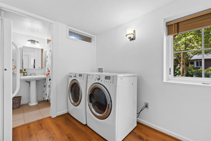 A bright laundry room with wooden floors features a front-loading washer and dryer side by side. A window lets in natural light. An open door leads to a bathroom with a pedestal sink and a shower curtain with colorful dots.