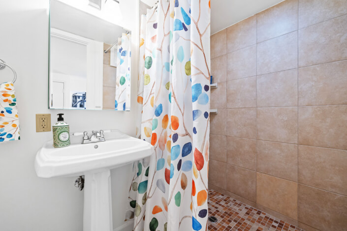 A bathroom with a pedestal sink, a mirror above it, and a soap dispenser. A colorful shower curtain with a leaf pattern separates the sink from a tiled, walk-in shower area with brown tiles and a mosaic tile floor.
