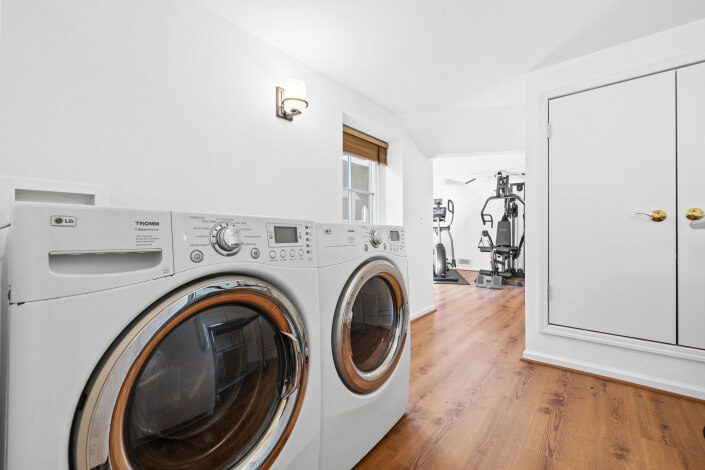 A laundry room with a front-loading washer and dryer side by side, wood flooring, a window with a brown shade, a wall light, and a view into a home gym with fitness equipment.